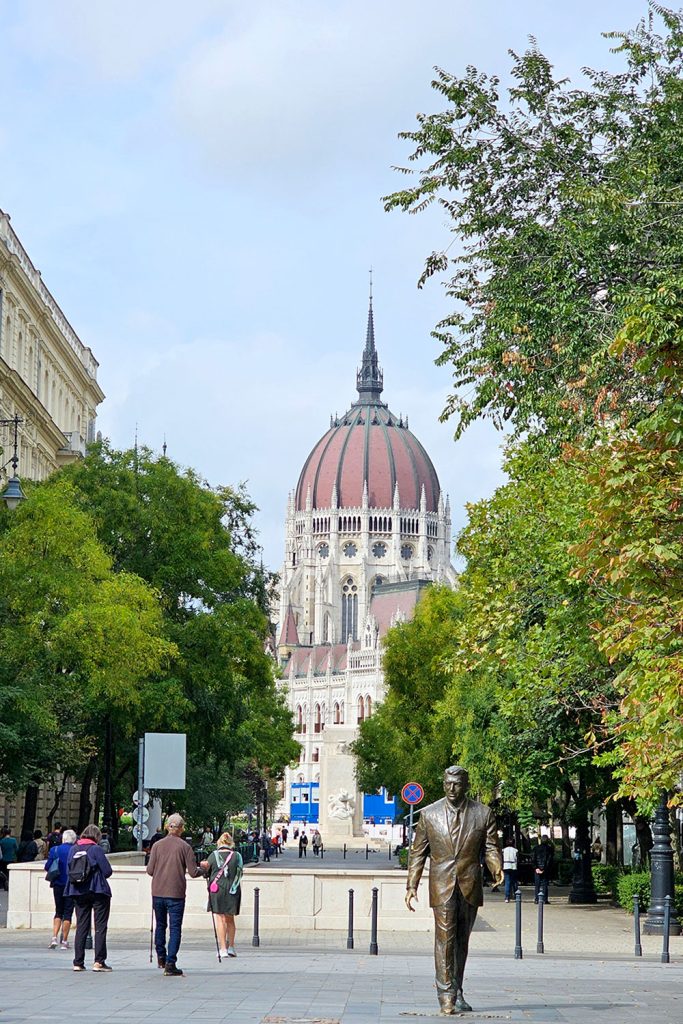 People dressed in light autumn layers walk through Liberty Square in Budapest on a typical fall day, with the Hungarian Parliament building and colorful foliage in the background.