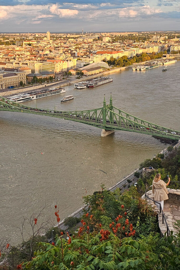 woman in a beige trench coat is taking photos of herself on a nice sunny autumn day at Gellért Hill, with the Liberty Bridge in the background